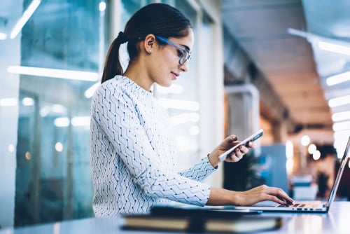 professional woman looking at device while working at laptop