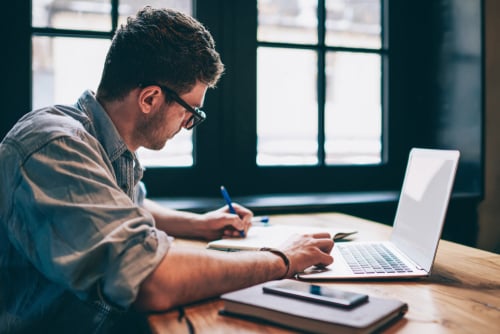 millenial male working at desk with open laptop and pen and paper