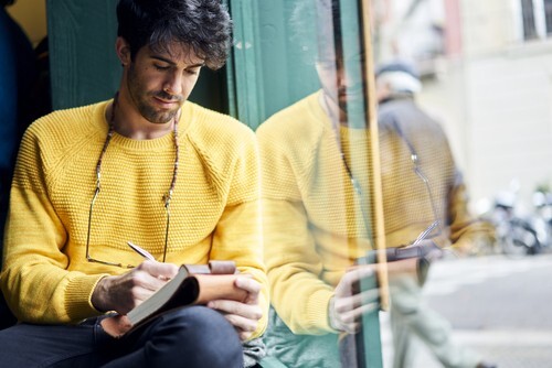 man writing in notebook next to glass window