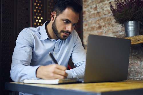 man writing notes while looking at computer screen